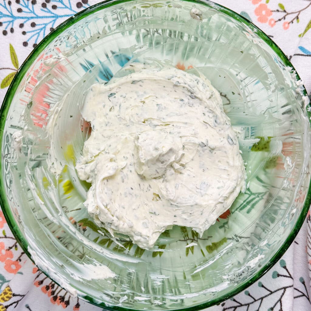 A glass bowl contains a creamy white mixture with visible green herb flecks—the perfect topping for This Crescent Dough Veggie Pizza. The bowl sits on a floral-patterned tablecloth.