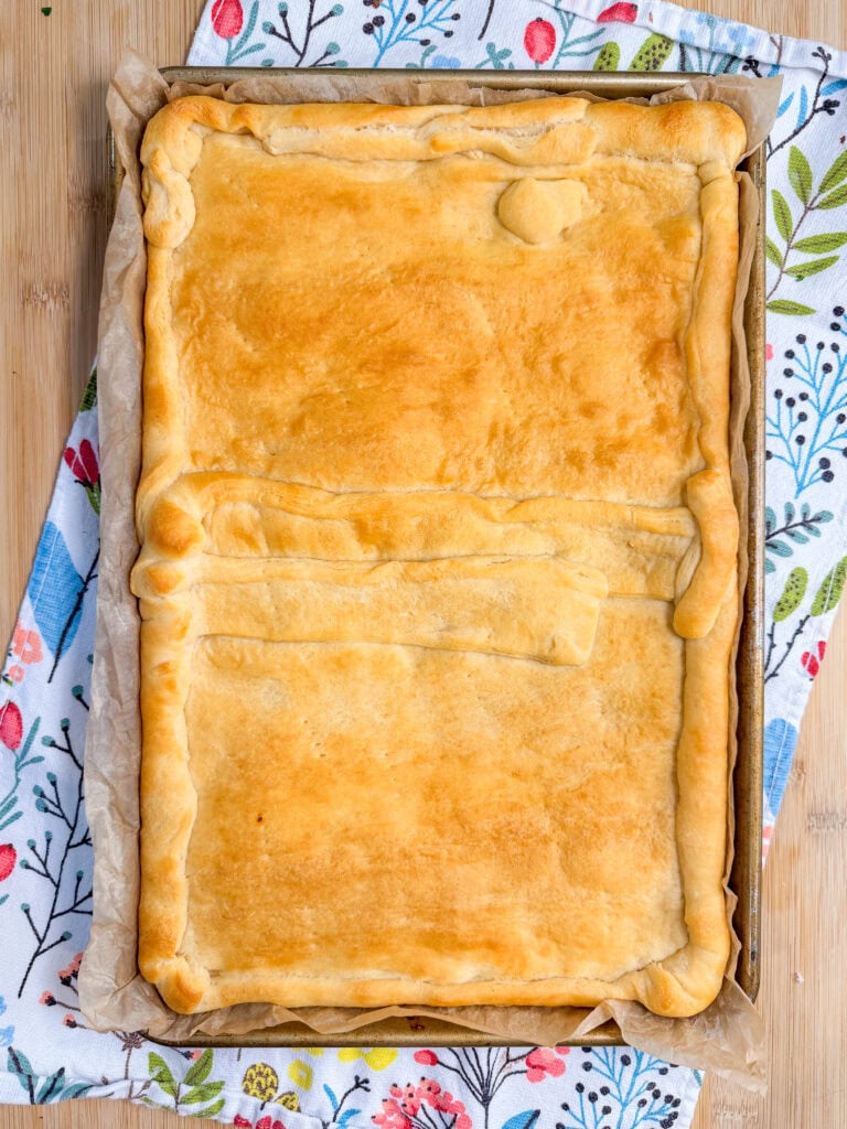 This Crescent Dough Veggie Pizza features a golden-brown rectangular pie with a slightly uneven crust, sitting on parchment paper atop a floral-patterned cloth, viewed from above.