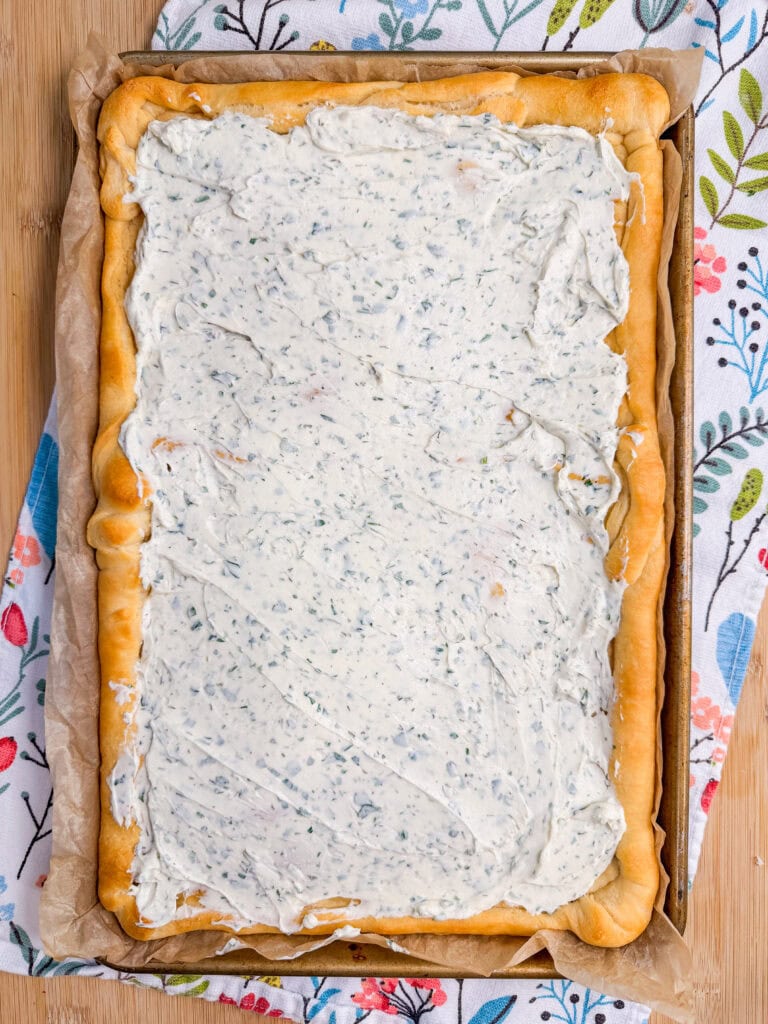 This Crescent Dough Veggie Pizza features a rectangular sheet of baked dough topped with a thick layer of creamy, herb-filled spread, resting on parchment paper over a floral-patterned cloth.