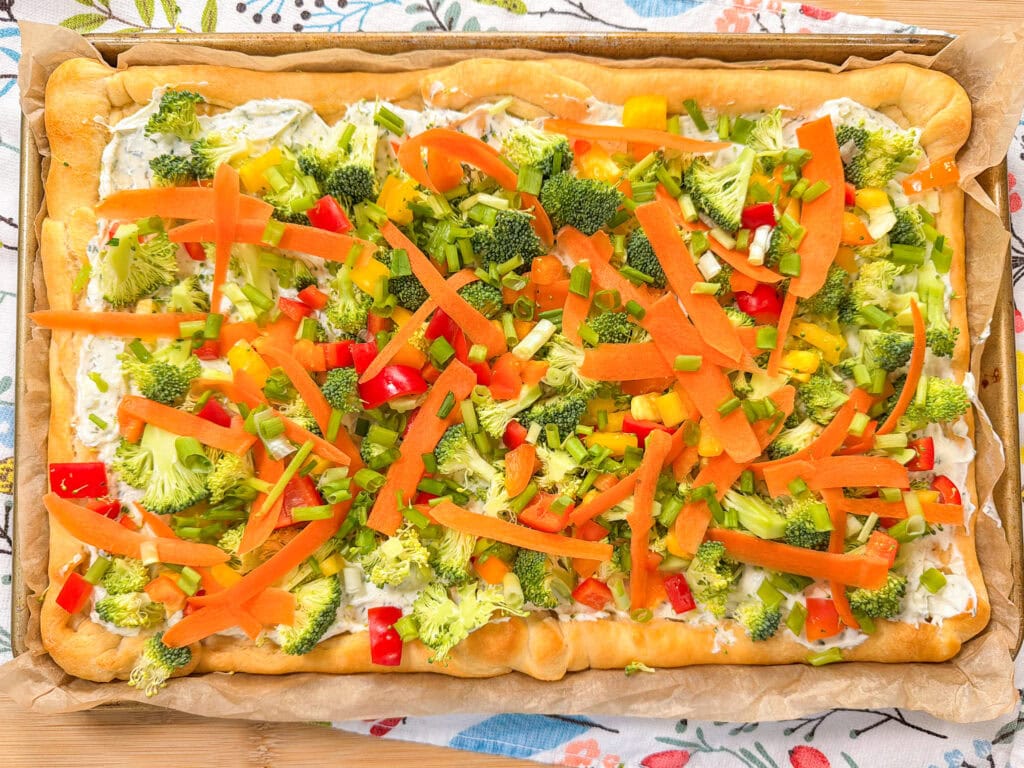 This Crescent Dough Veggie Pizza features a golden rectangular crust topped with cream cheese, chopped broccoli, red and yellow bell peppers, green onions, and thin carrot strips on a baking tray.