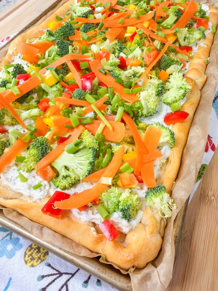 This Crescent Dough Veggie Pizza is baked on a sheet, topped with broccoli, carrots, bell peppers, green onions, and creamy white sauce over a golden crust. The image is bright and colorful, highlighting the fresh vegetables.