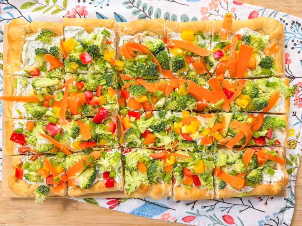 This Crescent Dough Veggie Pizza is a rectangular treat cut into squares, topped with broccoli, carrots, and bell peppers on a creamy spread, all served on a colorful floral-patterned cloth.