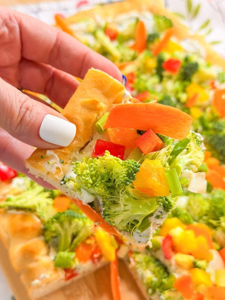 A hand with white nail polish holds a square slice of this Crescent Dough Veggie Pizza, topped with broccoli, carrots, red and yellow bell peppers, and green onions on a creamy base. The full pizza is visible in the background.