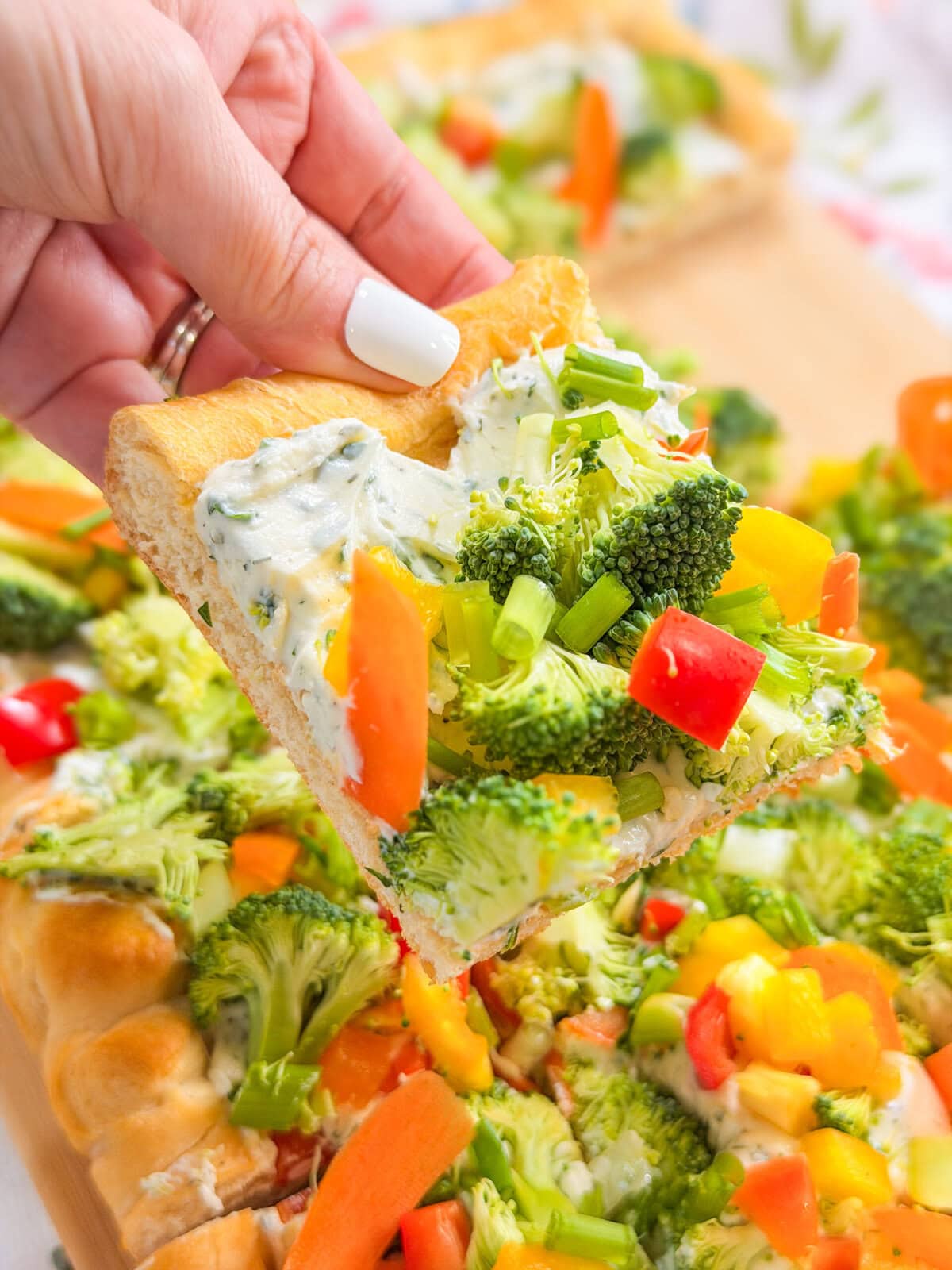 A hand holding a square slice of this Crescent Dough Veggie Pizza, topped with broccoli, carrots, bell peppers, green onions, and creamy white sauce on a thick, golden crust. Other slices are visible in the background.