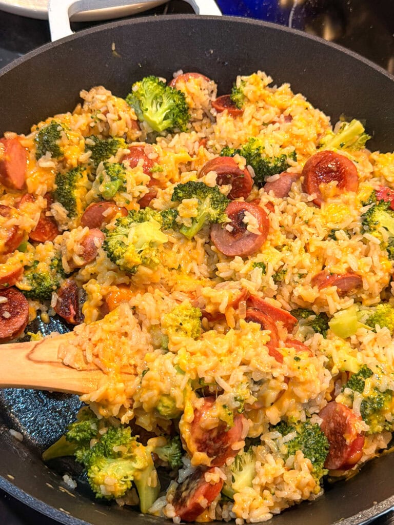 A skillet filled with a delicious Skillet Kielbasa with Rice—a savory mixture of rice, broccoli florets, sliced sausage, and melted cheese—being stirred with a wooden spatula.
