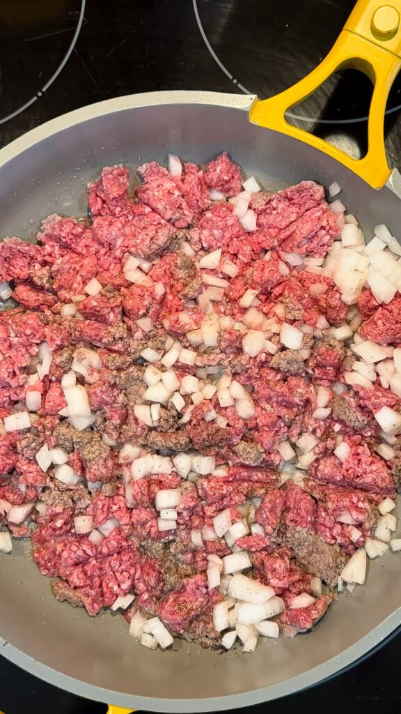 Raw ground beef and diced onions cooking in a skillet with yellow handles on a stovetopโthe perfect savory base for a flavorful Slow Cooker Stuffed Pepper Soup.