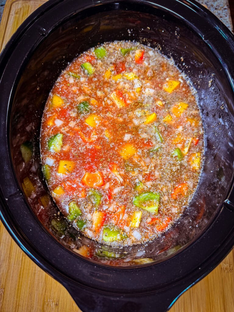 A slow cooker filled with a mixture of diced tomatoes, green and yellow bell peppers, onions, and seasonings in a broth, ready to be cookedโthis Slow Cooker Stuffed Pepper Soup is perfect for cozy evenings. The cooker sits on a wooden surface.
