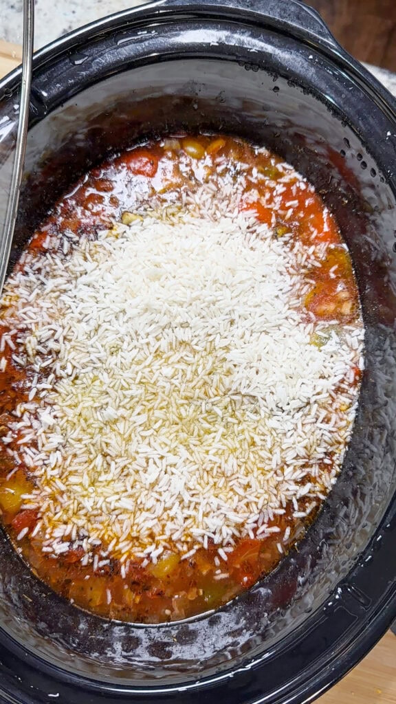 Uncooked white rice sits atop a mixture of vegetables and sauce in a black slow cooker, ready to be stirred and cooked for a comforting Slow Cooker Stuffed Pepper Soup.