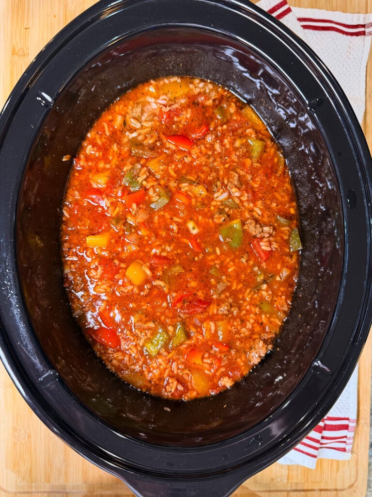A slow cooker filled with Slow Cooker Stuffed Pepper Soupโa chunky stew made of ground meat, rice, tomatoes, and chopped green and red bell peppers in a rich red sauce. The cooker sits on a wooden surface next to a red-and-white striped towel.