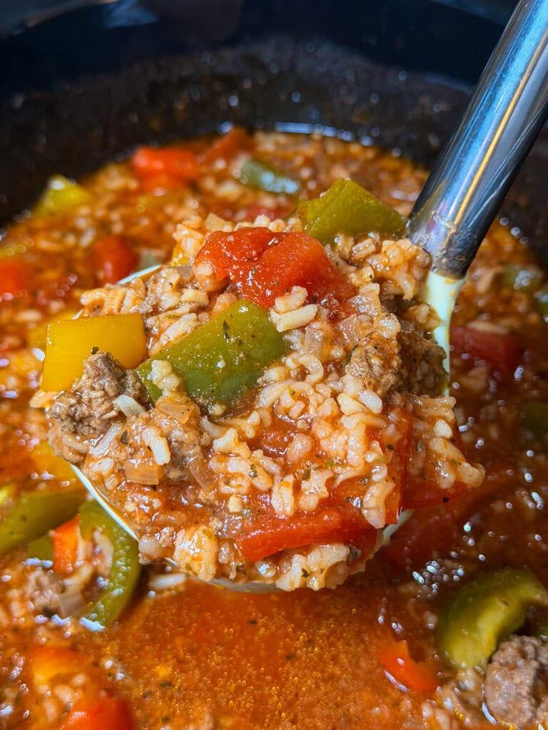 A close-up of a ladle filled with Slow Cooker Stuffed Pepper Soup, featuring ground beef, rice, tomatoes, and green and yellow bell peppers. The soup is chunky and colorful, with a rich tomato broth in the background.