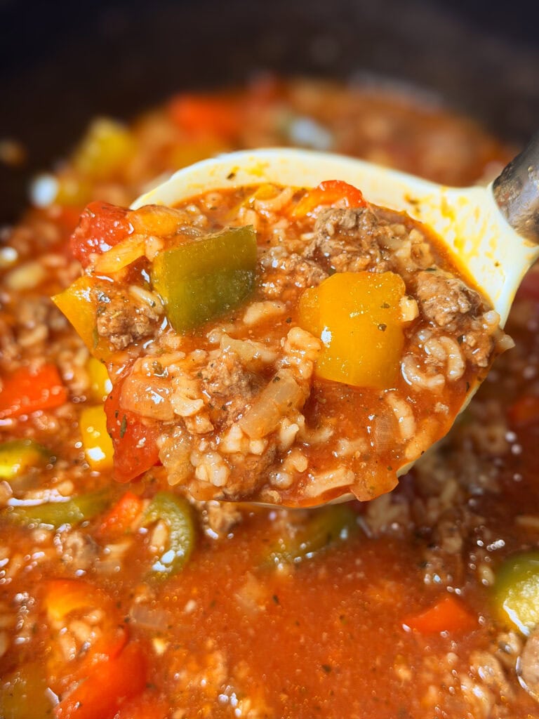 A close-up of a ladle holding Slow Cooker Stuffed Pepper Soup, featuring ground beef, rice, diced tomatoes, and chunks of green and yellow bell peppers in a rich tomato-based broth.