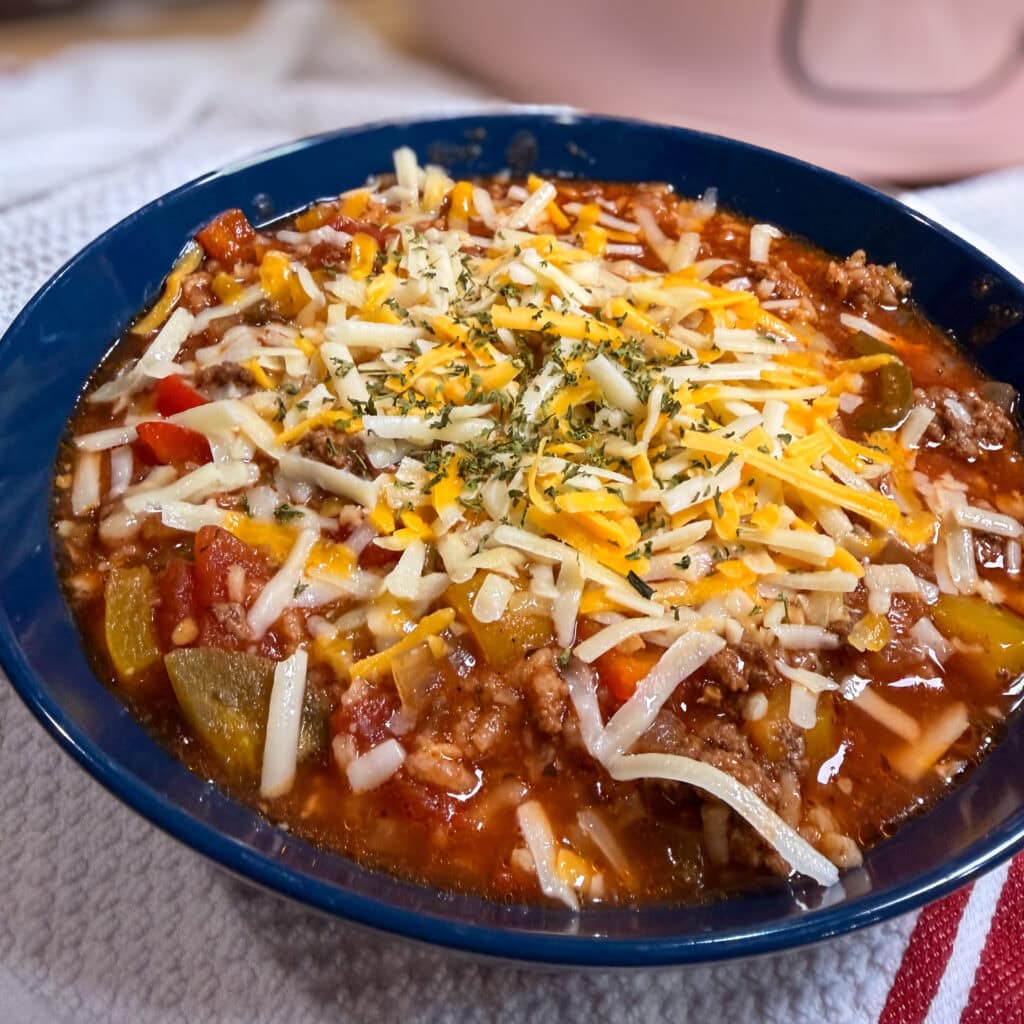A bowl of hearty chili reminiscent of Slow Cooker Stuffed Pepper Soup, topped with shredded cheese and chopped herbs, featuring ground meat, beans, tomatoes, and sliced peppers, served on a white textured surface.