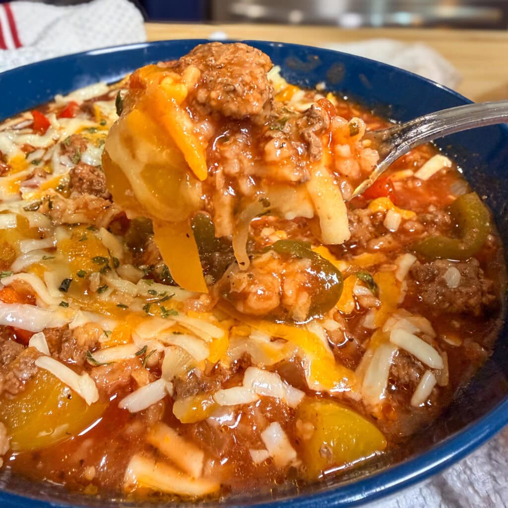 A close-up of a bowl of chunky Slow Cooker Stuffed Pepper Soup with ground beef, bell peppers, tomatoes, and melted shredded cheese, with a spoon lifting a hearty portion.