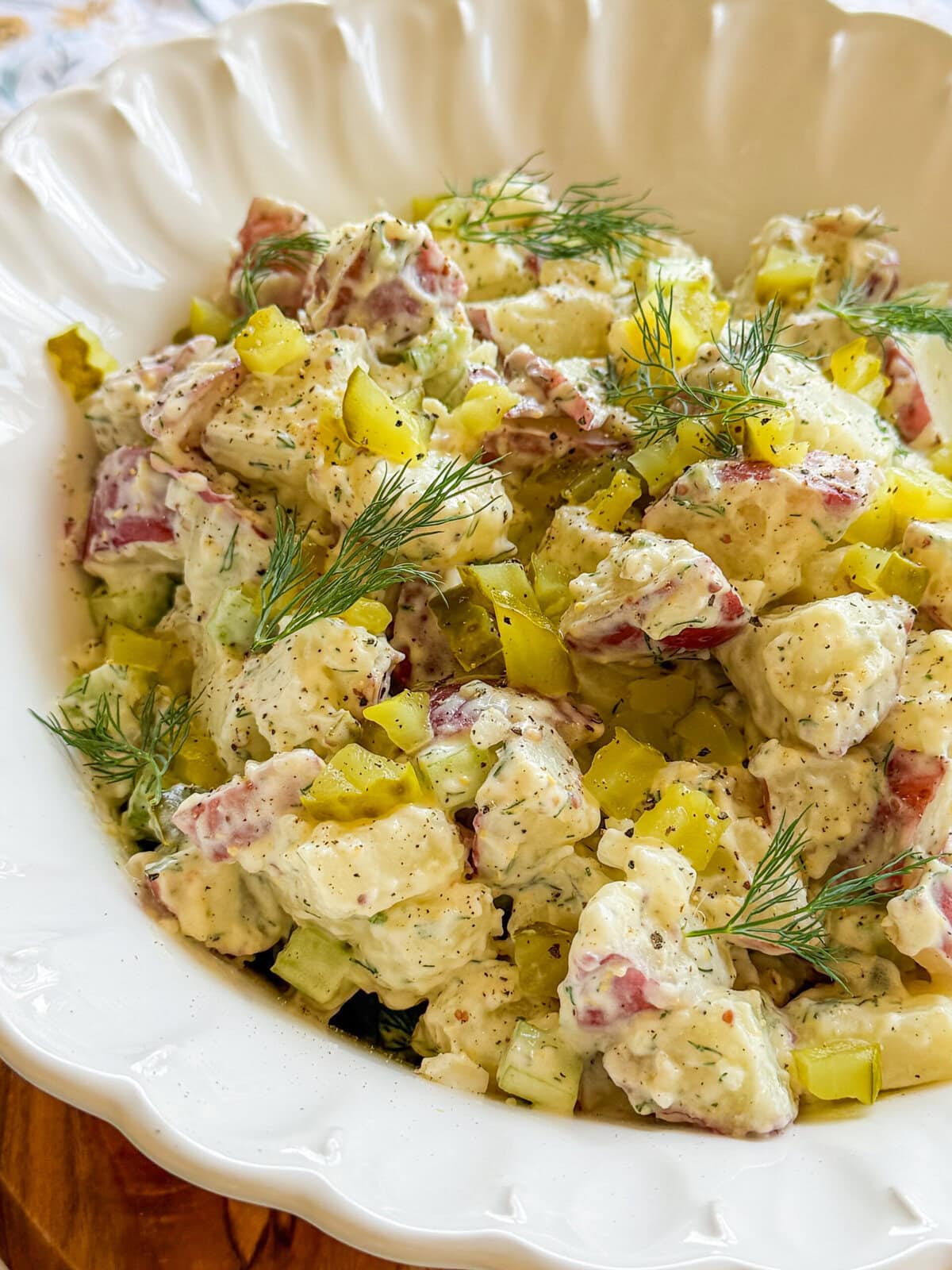 A close-up of creamy potato salad with diced red potatoes, chopped pickles, celery, and fresh dill is served in a white scalloped bowl—perfect alongside Fruity Pie Biscuit Bombs for a delightful spread.