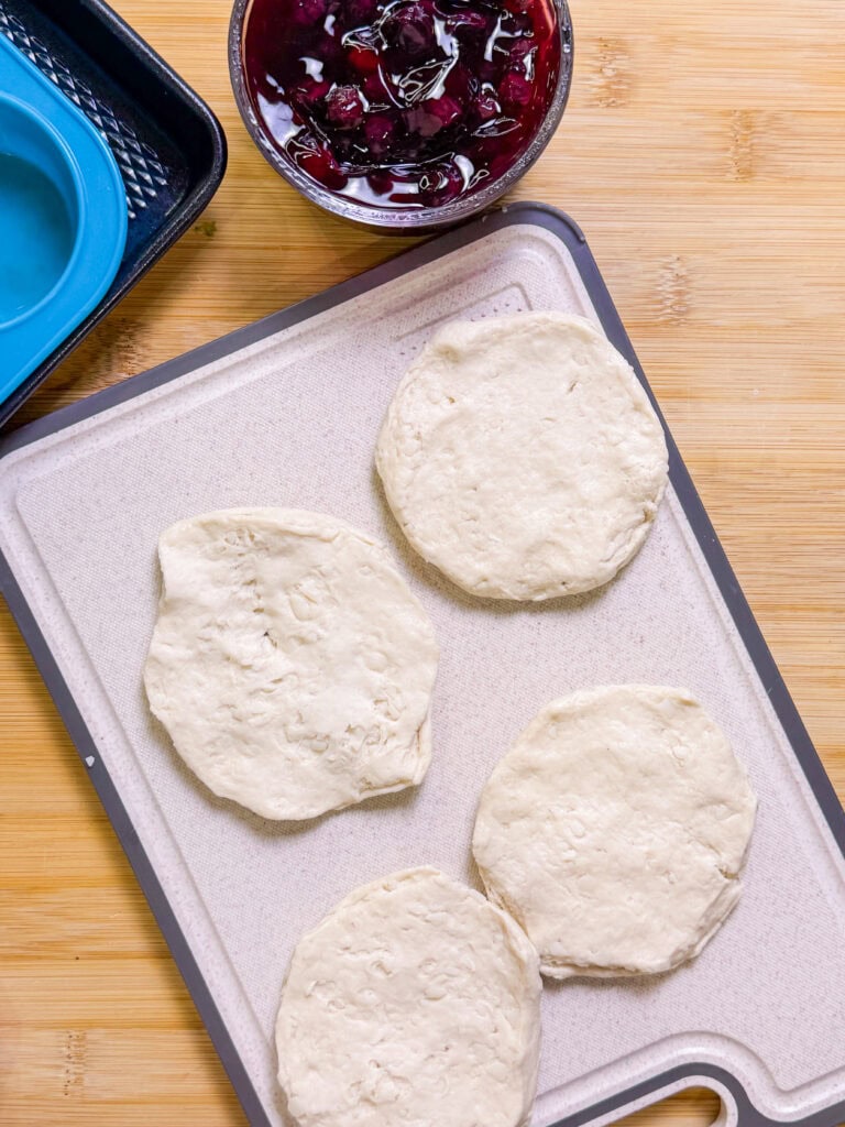 Four round pieces of raw biscuit dough are arranged on a cutting board next to a bowl of berry filling and a blue muffin tin, ready to be transformed into delicious Fruity Pie Biscuit Bombs on a wooden surface.