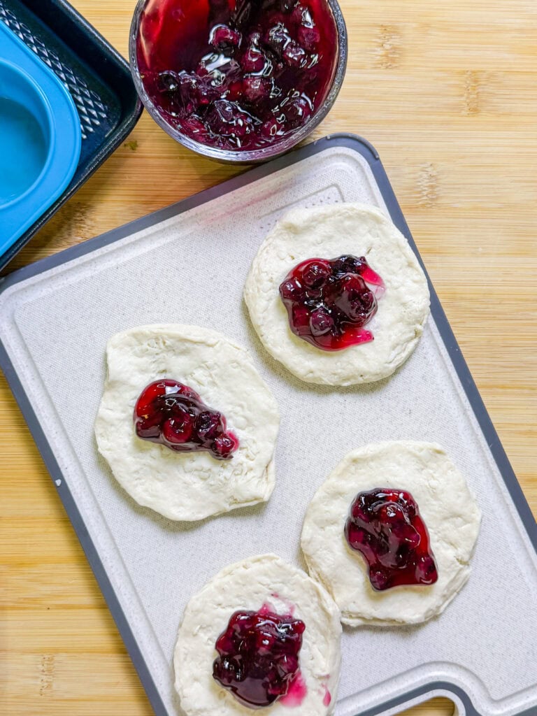 Four round Fruity Pie Biscuit Bombs sit on a cutting board, each topped with a spoonful of berry filling. Nearby are a bowl of berry filling and a blue muffin tin, all set on a wooden surface.