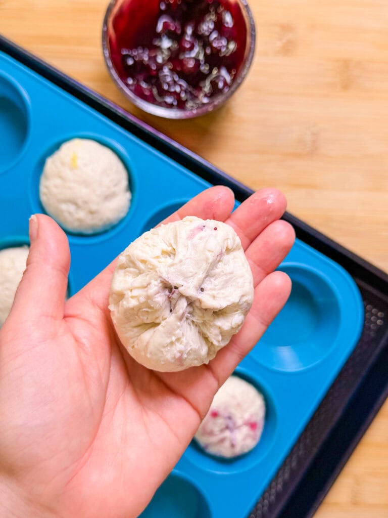 A hand holds an uncooked Fruity Pie Biscuit Bomb filled with jam above a blue silicone muffin tray. In the background, a bowl of dark berry jam sits on a wooden surface.
