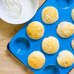 A blue silicone muffin tray holds six Fruity Pie Biscuit Bombs next to a white bowl of icing and a whisk, all placed on a wooden surface.