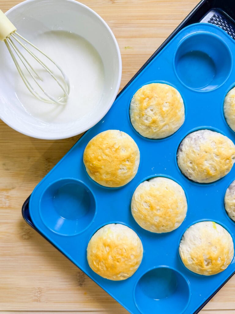 A blue silicone muffin tray holds six Fruity Pie Biscuit Bombs next to a white bowl of icing and a whisk, all placed on a wooden surface.
