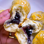 A hand holds a Fruity Pie Biscuit Bomb broken in half, revealing a gooey purple berry filling inside. More round, golden-brown pastries are visible on a plate in the background.