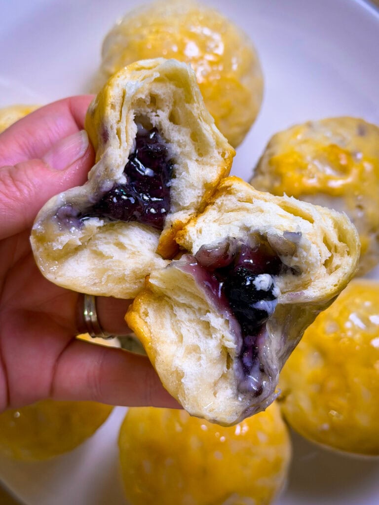A hand holds a Fruity Pie Biscuit Bomb broken in half, revealing a gooey purple berry filling inside. More round, golden-brown pastries are visible on a plate in the background.