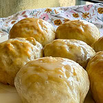 Close-up of several homemade glazed cinnamon rolls on a white plate, with a floral-patterned cloth in the background. The golden-brown rolls are covered in a shiny layer of icing, resembling the irresistible appeal of Fruity Pie Biscuit Bombs.