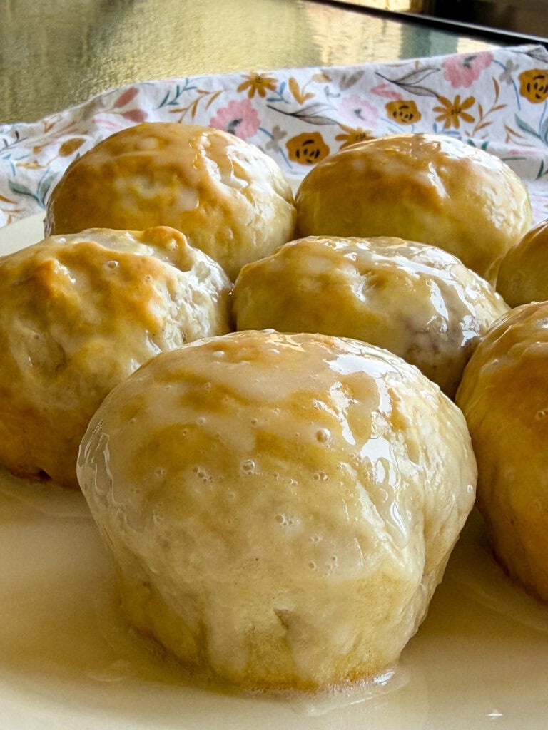 Close-up of several homemade glazed cinnamon rolls on a white plate, with a floral-patterned cloth in the background. The golden-brown rolls are covered in a shiny layer of icing, resembling the irresistible appeal of Fruity Pie Biscuit Bombs.