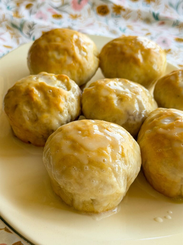 Seven golden-brown Fruity Pie Biscuit Bombs, covered in a glossy glaze, are arranged on a cream plate, with a floral-patterned tablecloth in the background.