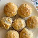 Seven Fruity Pie Biscuit Bombs are arranged on a white plate, each covered in shiny icing. The plate sits on a surface adorned with a floral patterned tablecloth.
