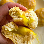 A hand holds a soft, fluffy bun torn open to reveal a gooey, yellow custard filling—reminiscent of Fruity Pie Biscuit Bombs—with more buns stacked invitingly on a plate in the background.
