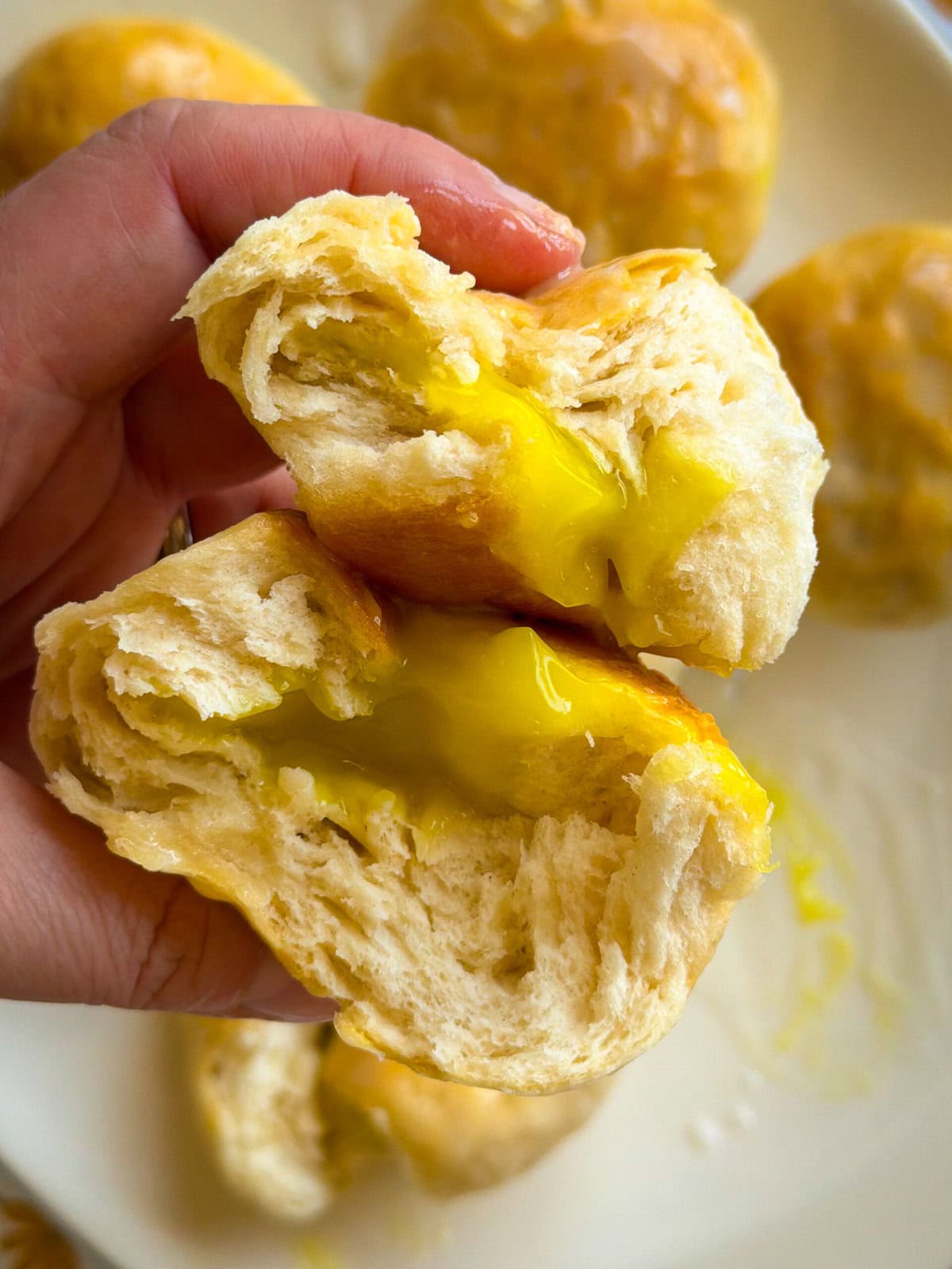 A hand holds a soft, fluffy bun torn open to reveal a gooey, yellow custard filling—reminiscent of Fruity Pie Biscuit Bombs—with more buns stacked invitingly on a plate in the background.