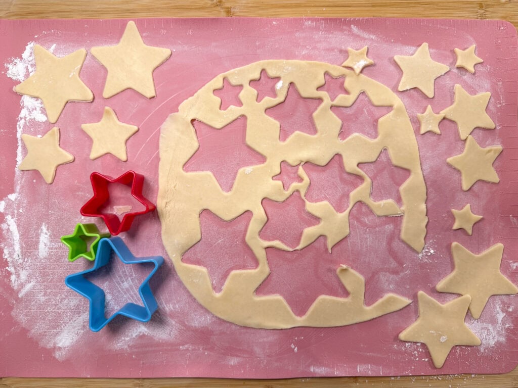 Star-shaped cookie dough cutouts and colorful star-shaped cookie cutters are arranged on a pink floured surface, with leftover dough showing star-shaped gaps—perfect for topping a Cherry Slab Pie with delightful, whimsical flair.