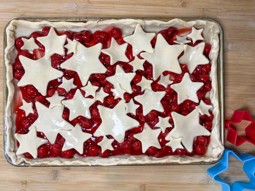 A Cherry Slab Pie with a crust decorated in star-shaped dough cutouts sits on a wooden surface. Red and blue star-shaped cookie cutters are arranged to the right of the pie.