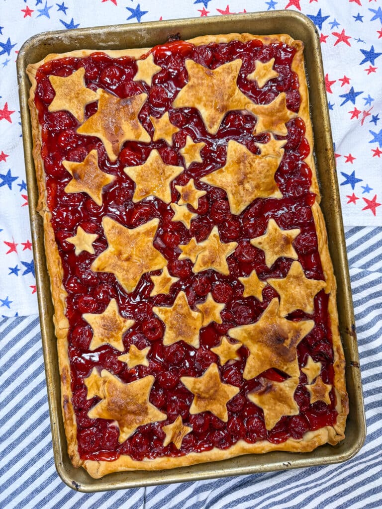 A Cherry Slab Pie with a golden crust, decorated with star-shaped pastry pieces on top, sits on a baking sheet atop a festive, star-patterned cloth background.