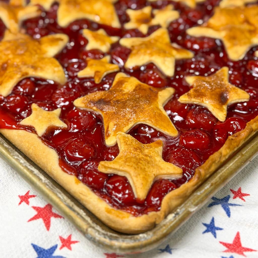 A rectangular Cherry Slab Pie topped with golden star-shaped crust pieces sits on a baking sheet, resting on a white cloth decorated with red and blue stars.
