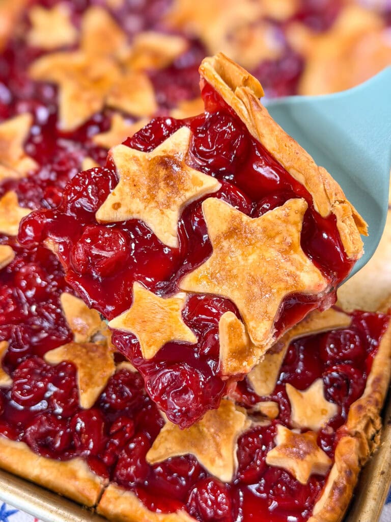 A close-up of a Cherry Slab Pie slice with golden, star-shaped pieces of crust on top, being lifted from the rest of the pie with a spatula. The cherry filling is glossy and vibrant red.