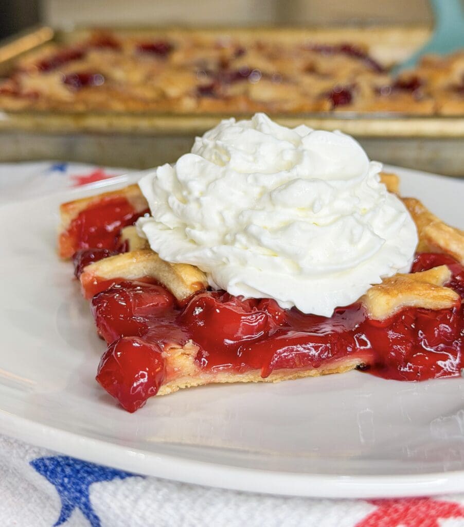 A slice of Cherry Slab Pie with a lattice crust is topped with a generous swirl of whipped cream on a white plate. The vibrant red, glossy filling stands out, with more pie tempting in the background.