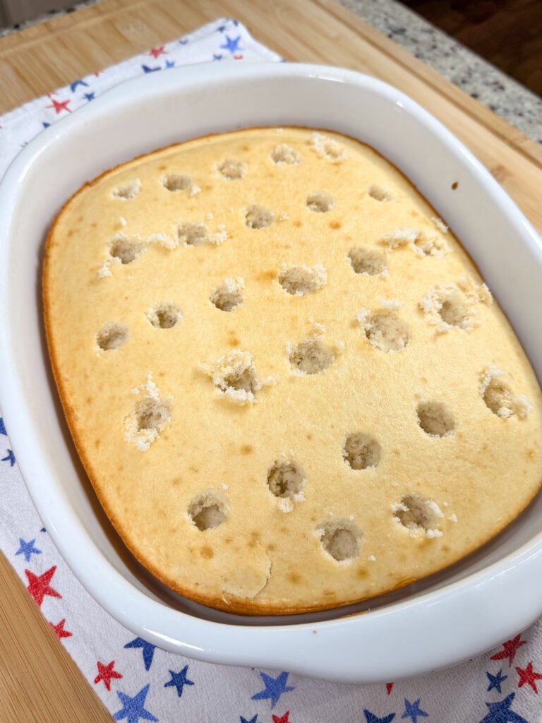 A Patriotic Jell-O Poke Cake in a white dish features evenly spaced holes poked in the top, sitting on a white towel decorated with red and blue stars.