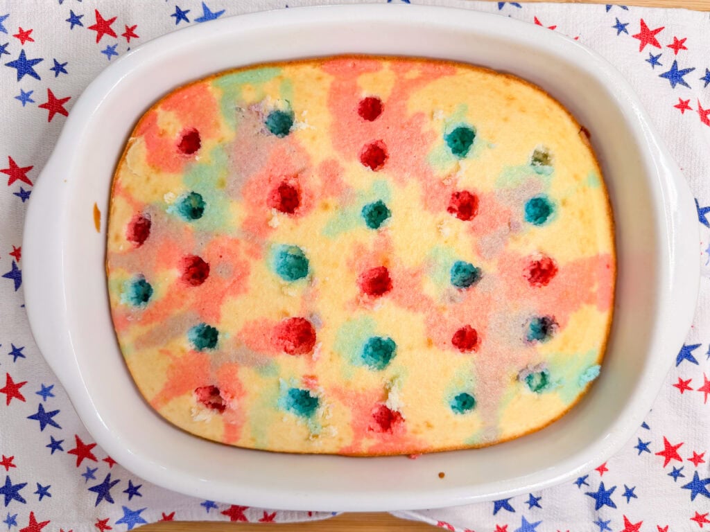 A Patriotic Jell-O Poke Cake in a white dish, featuring red and blue spots and swirls. It rests on a white towel decorated with red and blue stars.