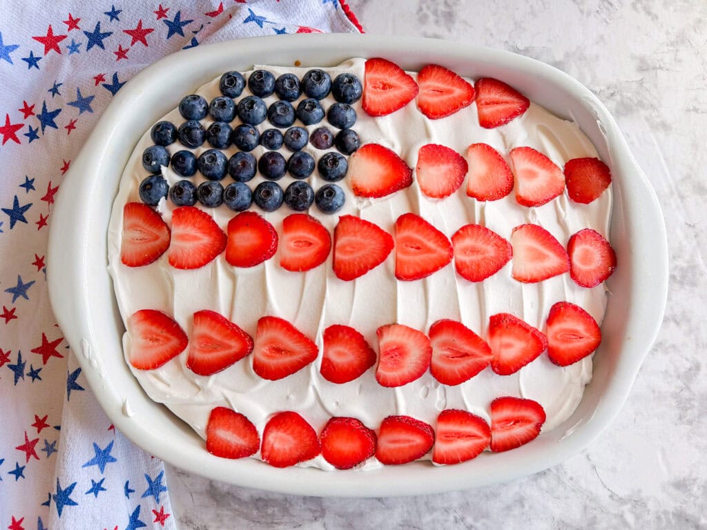 A rectangular Patriotic Jell-O Poke Cake topped with whipped cream, sliced strawberries in stripes, and blueberries in the corner to resemble the American flag, set on a surface beside a star-patterned cloth.