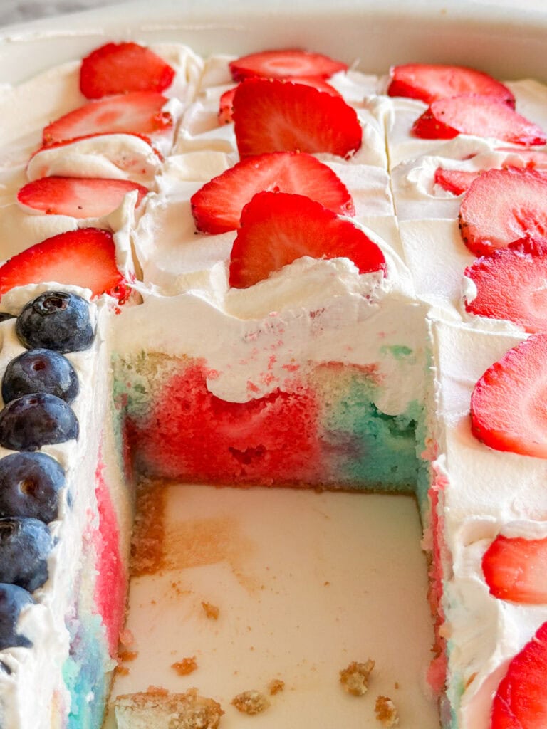 A close-up of a Patriotic Jell-O Poke Cake with red, white, and blue layers, topped with whipped cream, sliced strawberries, and blueberries. A section has been cut out, revealing the vibrant inside.