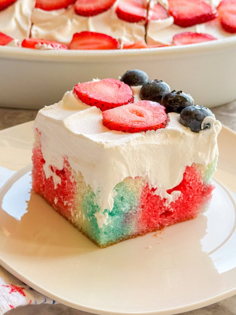 A square slice of Patriotic Jell-O Poke Cake topped with whipped cream, fresh strawberry slices, and blueberries sits on a white plate, with the rest of this festive cake in the background.