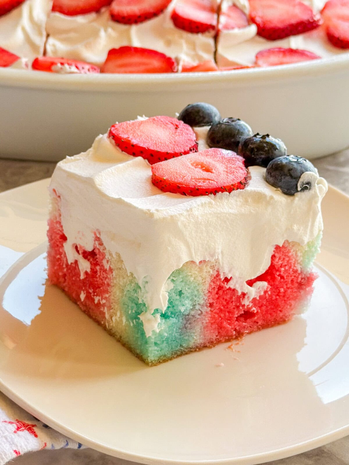 A square slice of Patriotic Jell-O Poke Cake topped with whipped cream, fresh strawberry slices, and blueberries sits on a white plate, with the rest of this festive cake in the background.