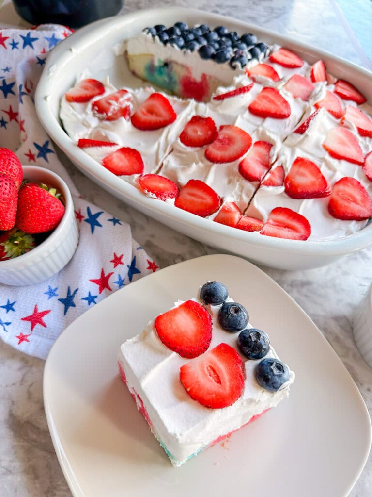 A Patriotic Jell-O Poke Cake topped with whipped cream, sliced strawberries, and blueberries is cut into squares. One square is served on a plate; the rest remains in a baking dish, with a bowl of strawberries nearby.