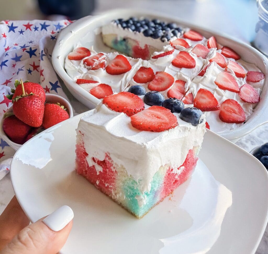 A slice of Patriotic Jell-O Poke Cake on a white plate, topped with whipped cream, fresh strawberries, and blueberries. The cake’s red, white, and blue layers stand out against the background featuring the whole festive cake and a bowl of strawberries.