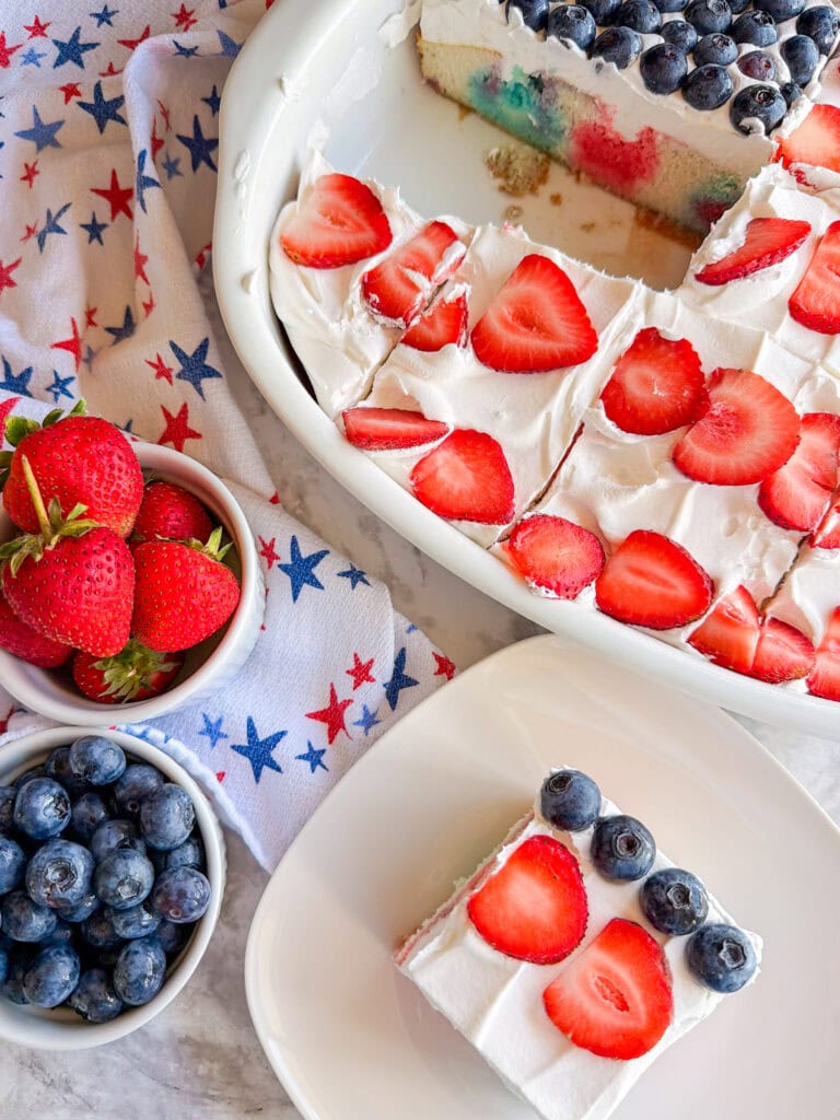 A tray of Patriotic Jell-O Poke Cake topped with whipped cream, sliced strawberries, and blueberries sits next to a star-patterned cloth, with bowls of fresh strawberries and blueberries, and a plated slice of cake in the foreground.