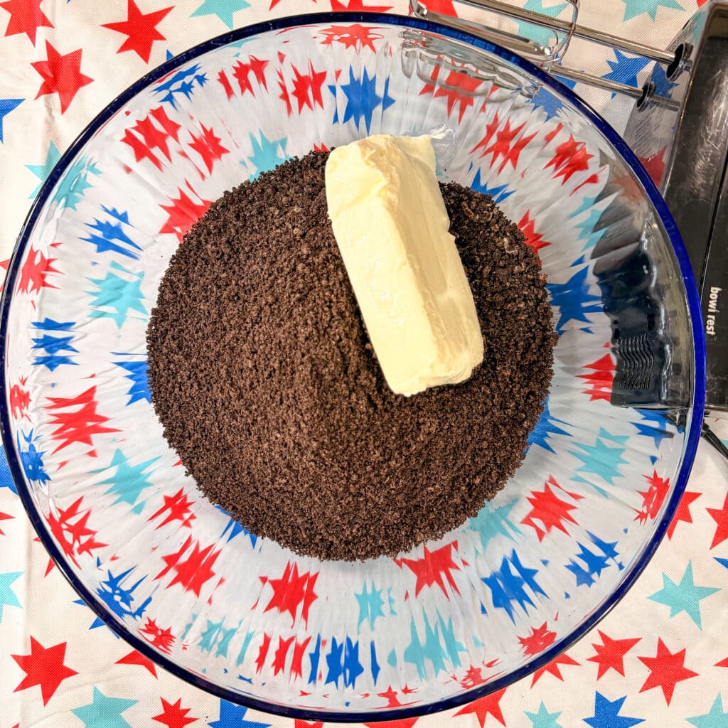 A large glass bowl contains a stick of butter atop a mound of finely crushed chocolate cookies, perfect for making Patriotic Oreo Truffles. The bowl rests on a festive tablecloth with red, blue, and light blue stars.