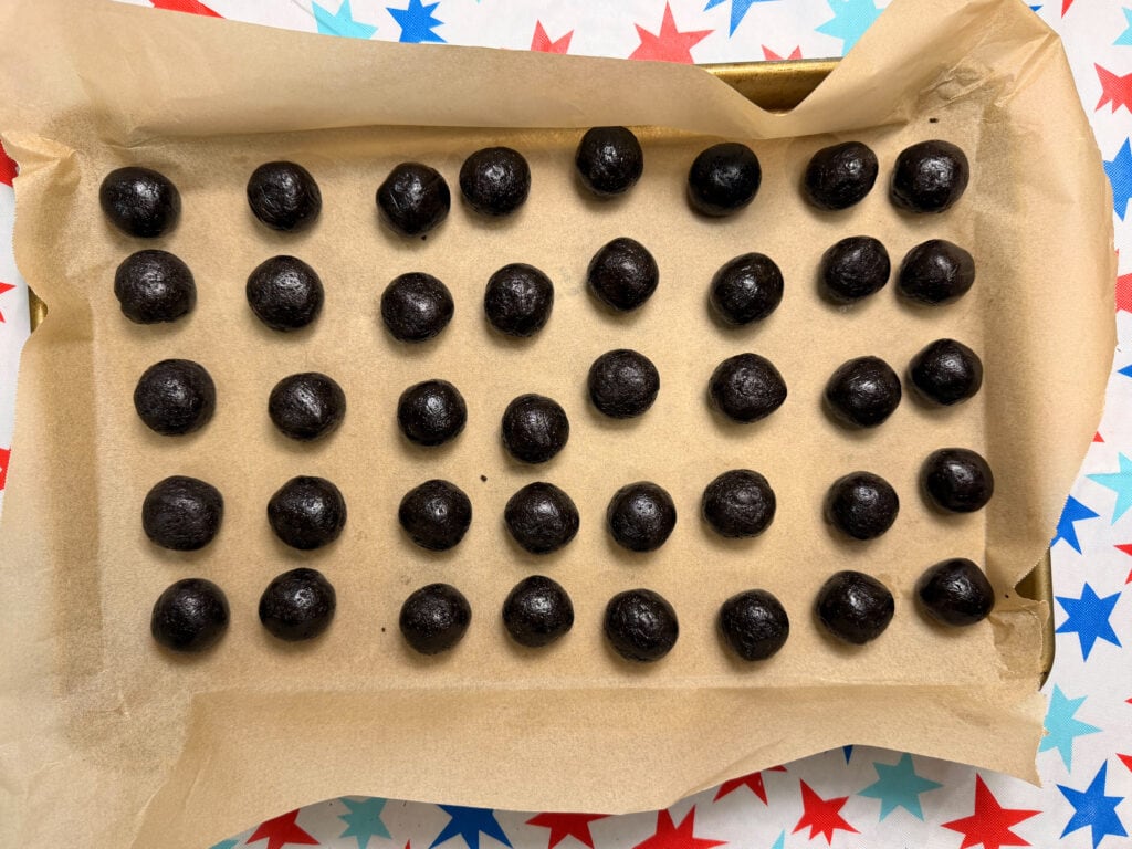 A parchment-lined baking tray holds five rows of evenly spaced Patriotic Oreo Truffles. The tray sits on a festive surface decorated with red, blue, and teal stars.