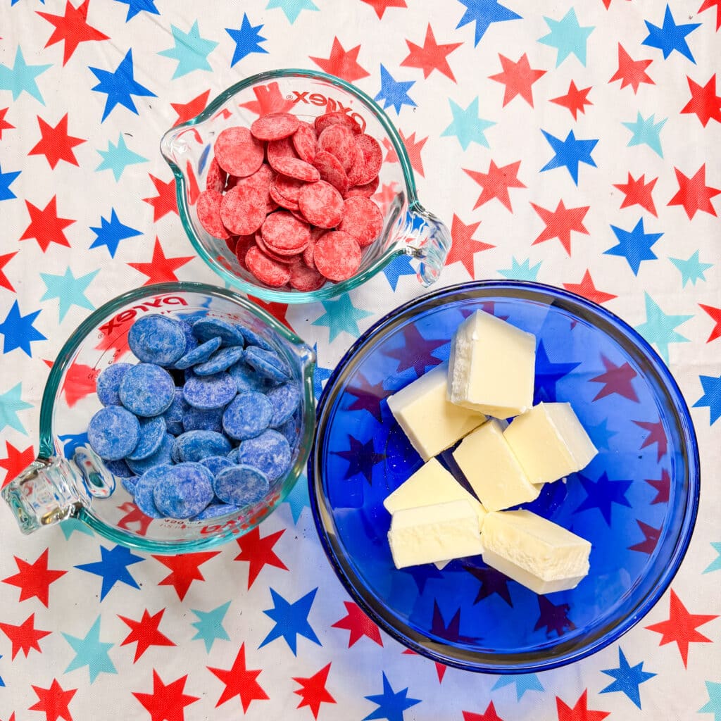 Three bowls on a red, white, and blue star-patterned surface: one with red candy melts, one with blue candy melts, and one with white chocolate or candy coating—perfect for making festive Patriotic Oreo Truffles.