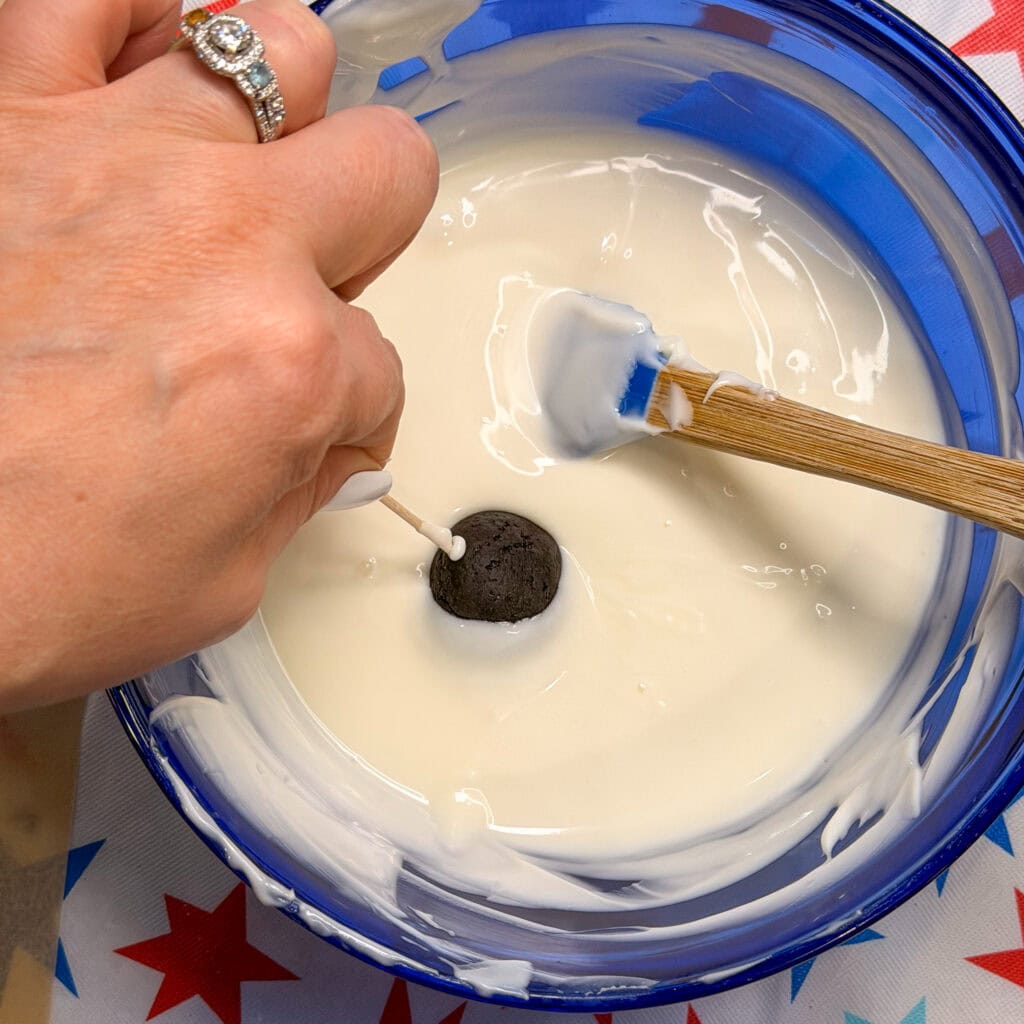 A hand holds a stick with a Patriotic Oreo Truffle being dipped into a bowl of melted white chocolate, using a wooden spatula to assist. The bowl rests on a star-patterned tablecloth.